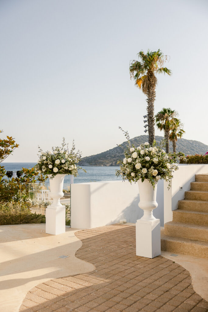 White floral arrangements in urns on pedestals by curved brick path and steps near water.