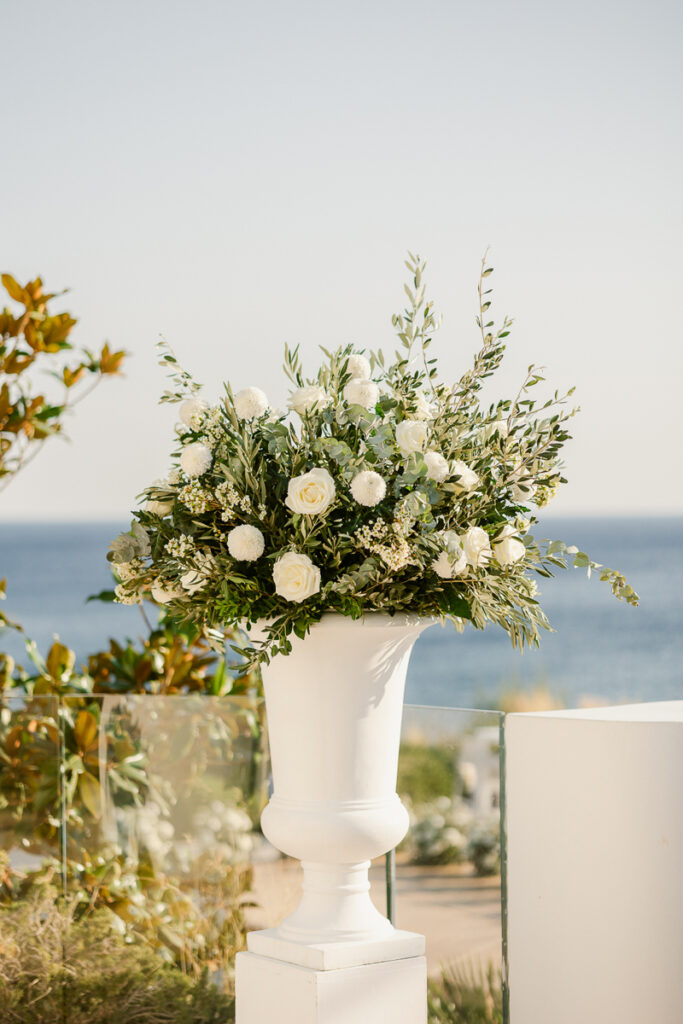 White urn with white flowers and green foliage by sea on a glass-railed terrace.