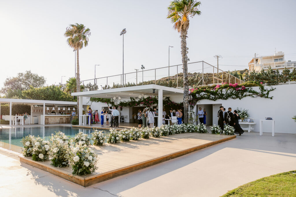 Outdoor venue with pool, floral decorations, a bar, palm trees, and people socializing under a white pavilion.