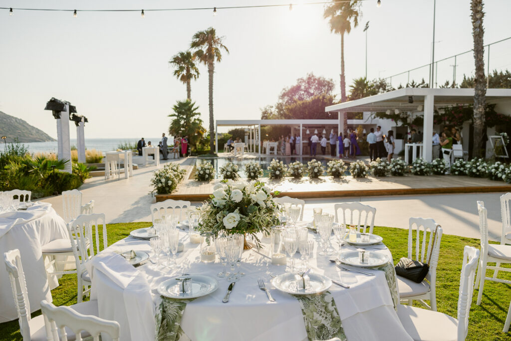 Outdoor event with tables set with white linens and floral centerpieces near a pool and ocean view.