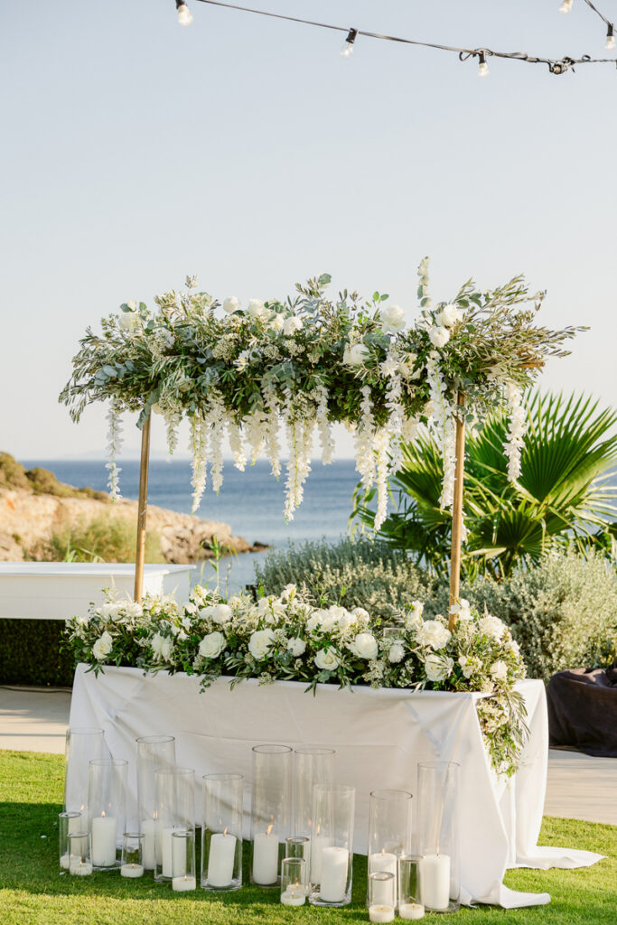 Outdoor table with white floral arrangement, cascading flowers, candles in glass holders, and string lights.