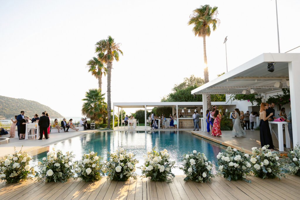 Outdoor poolside social event with guests, white floral decorations, palm trees, and hillside view.