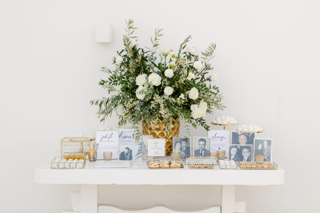 White table with large floral centerpiece, black-and-white photos, treats, pens, and calligraphy signs.