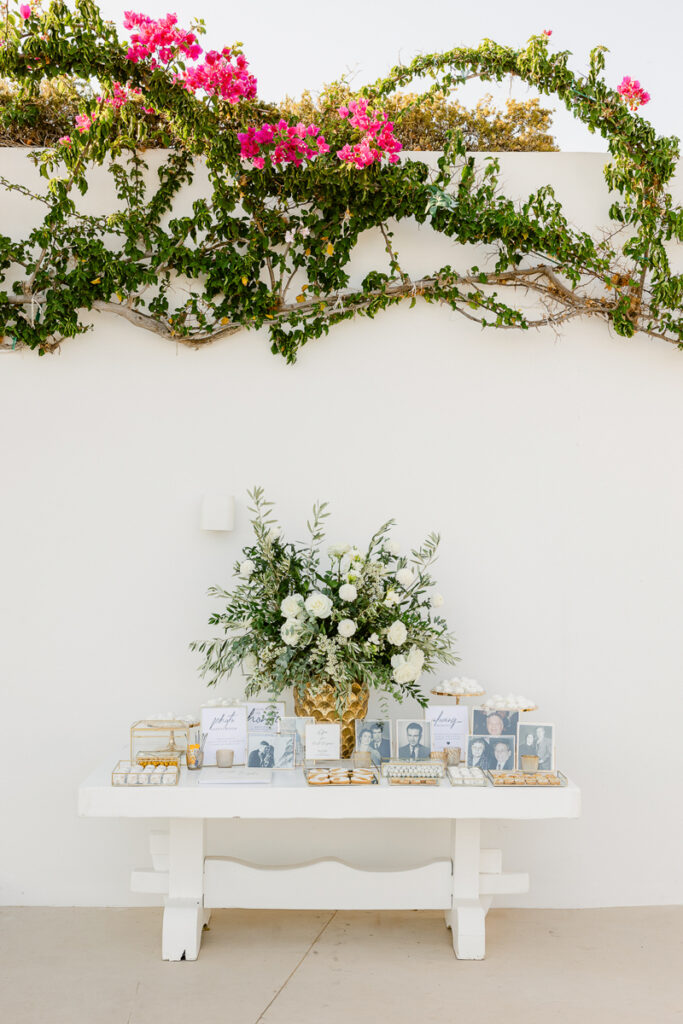 White table with a gold vase of white flowers, framed photos, and trays of sweets against a white wall with green vines and pink flowers.