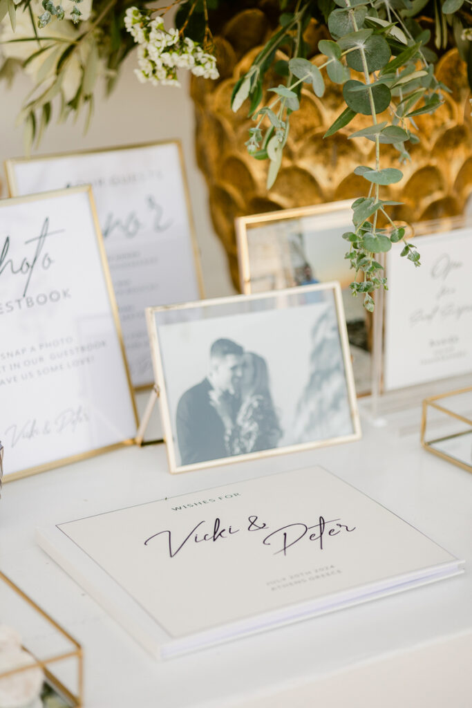 Wedding guestbook with names Vicki and Peter on white table with photos and greenery above.