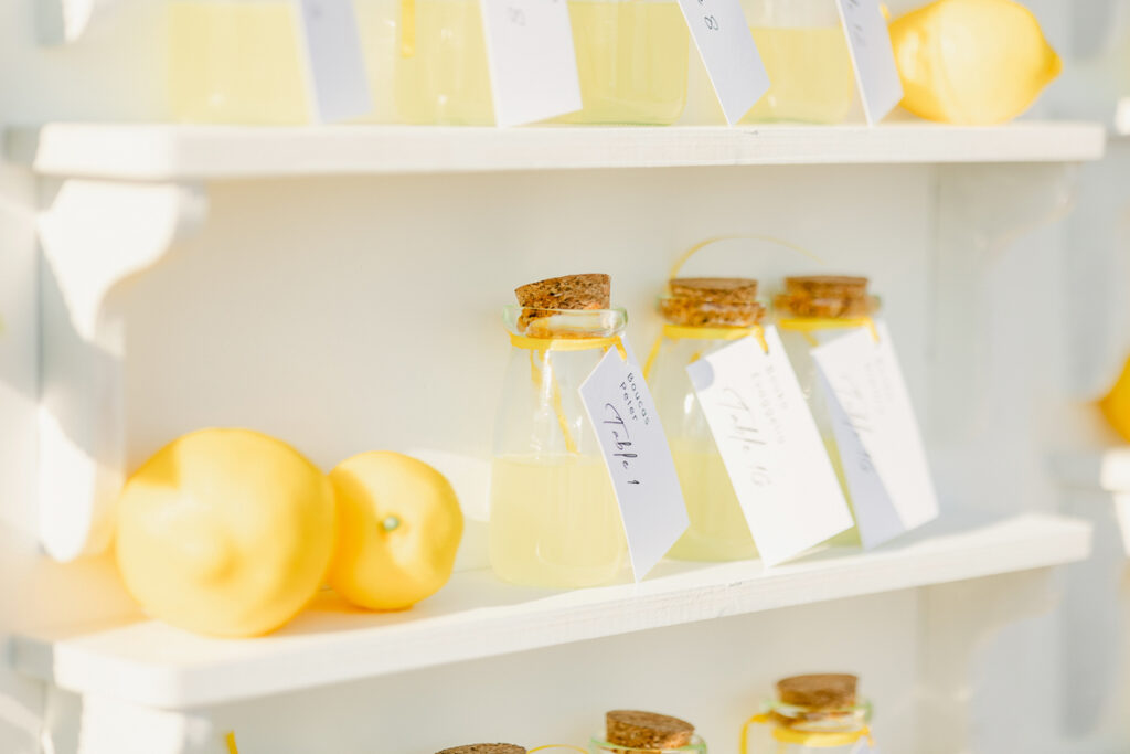 Glass bottles with cork stoppers filled with yellow liquid and whole lemons on white shelves.