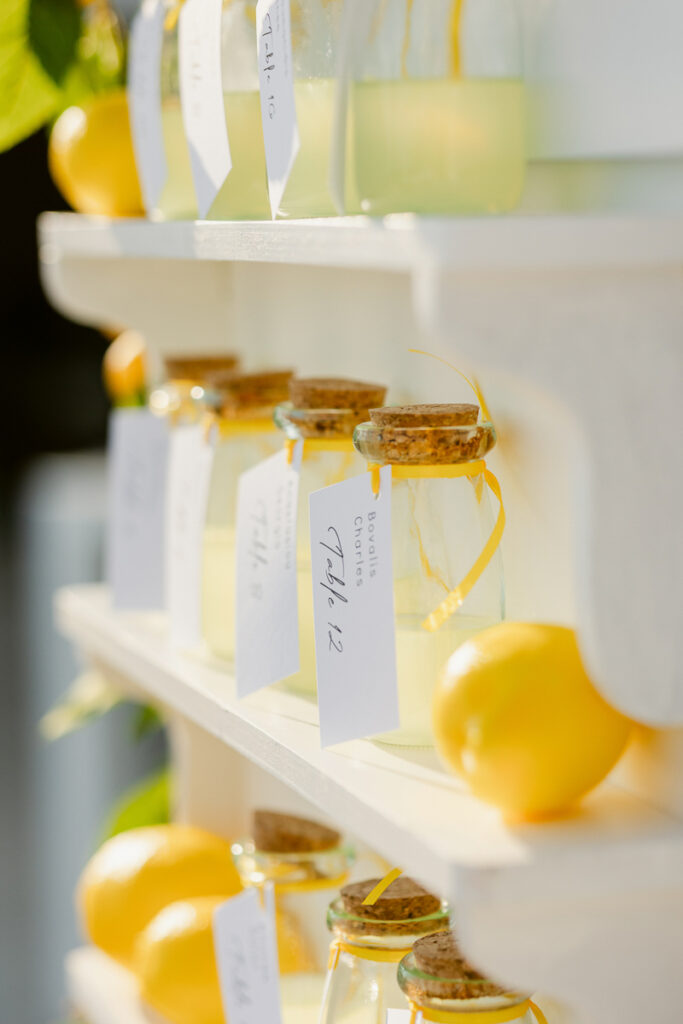 Glass jars with corks and tags holding yellow liquid arranged on white shelves with whole lemons.