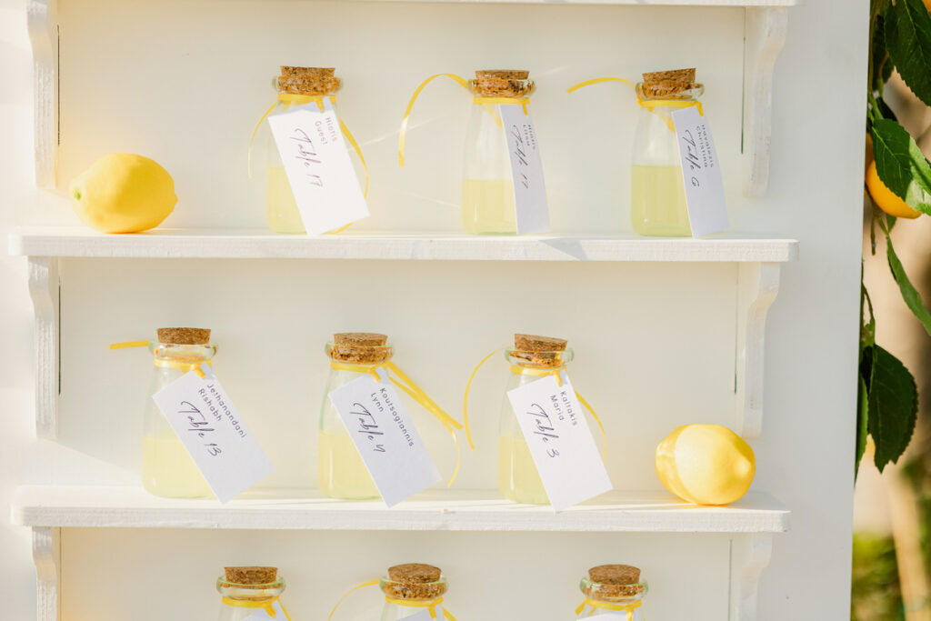 Small glass bottles with yellow liquid and tags on a white three-tiered shelf with lemons and foliage.