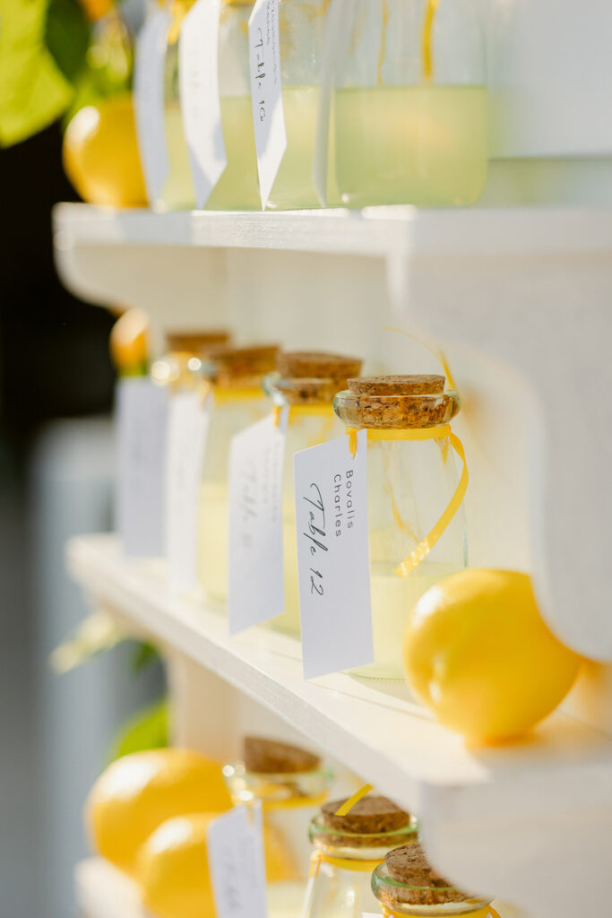 Glass bottles with corks containing pale yellow liquid and labeled tags on white shelves with lemons.