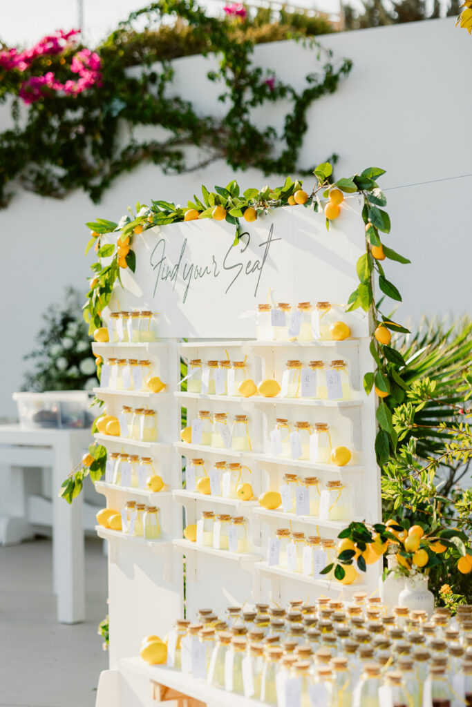 White seating display with glass bottles, corks, tags, lemon decor, and greenery outdoors.