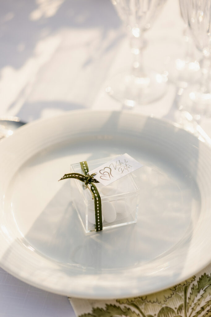 Clear box with white almonds tied with green ribbon and tag on a white plate at a formal setting