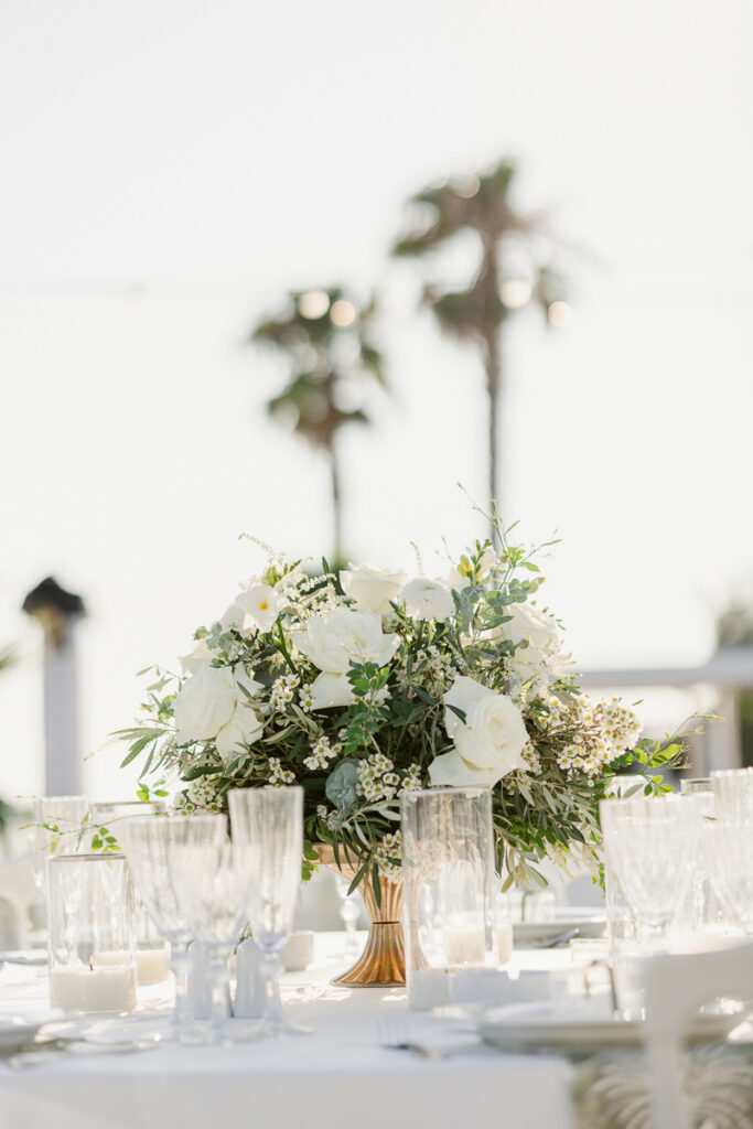 White rose and greenery centerpiece in gold vase on table set with glassware and plates, palm trees in background