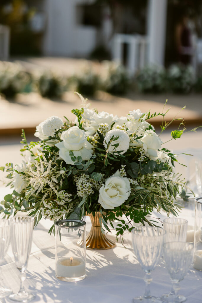 Floral centerpiece with white roses and greenery in a gold vase on a white tablecloth.