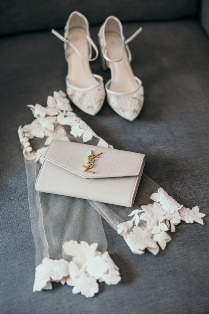 White high-heeled shoes, designer purse with gold logo, and sheer fabric with floral appliqué on gray surface