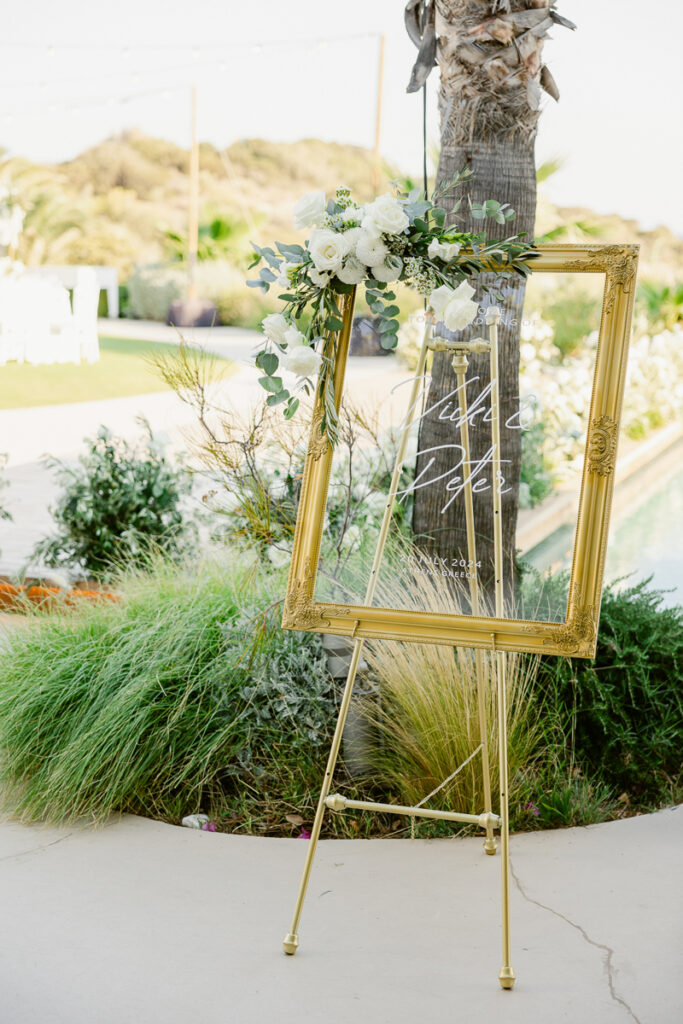 Gold-framed transparent wedding welcome sign with white floral decoration on gold easel outdoors