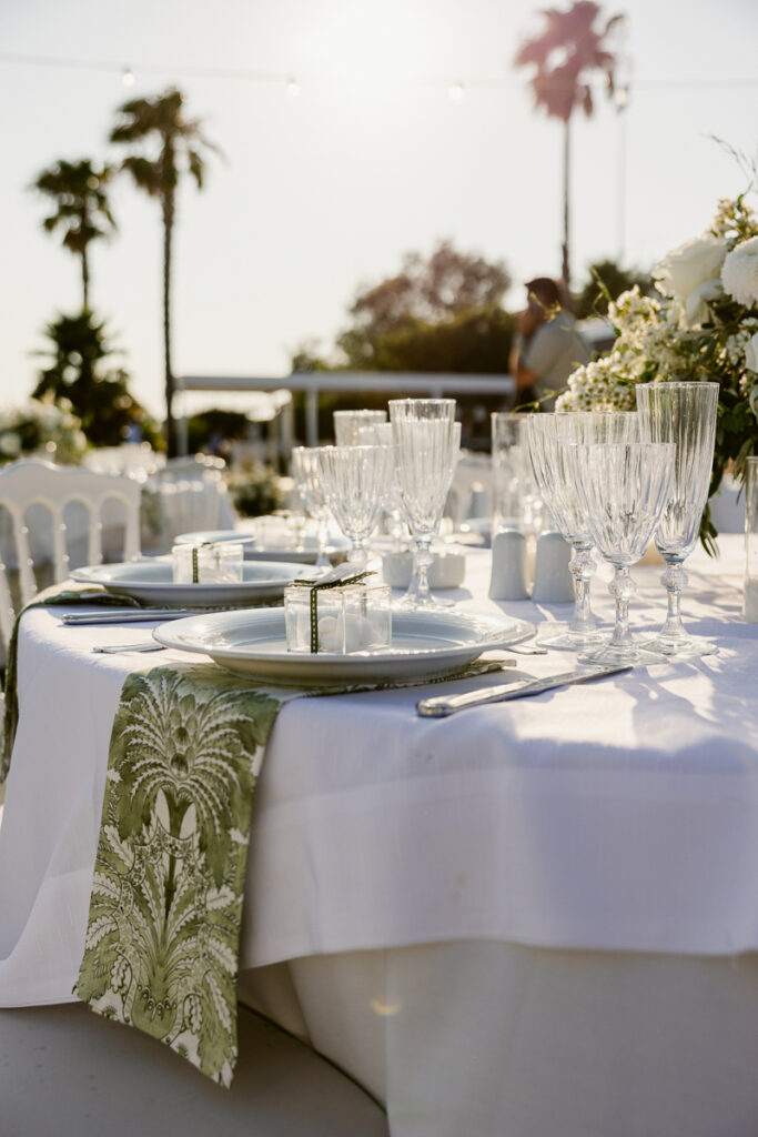 Outdoor dining table with white tablecloth, glassware, plates, green napkins, and floral centerpiece.
