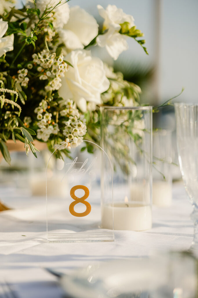 Clear acrylic table number 8 sign with white flowers, lit candle, and glassware on white tablecloth