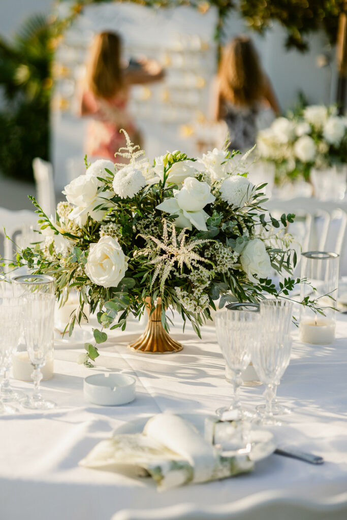 White floral centerpiece in gold vase on a table with glassware and candles at an event.