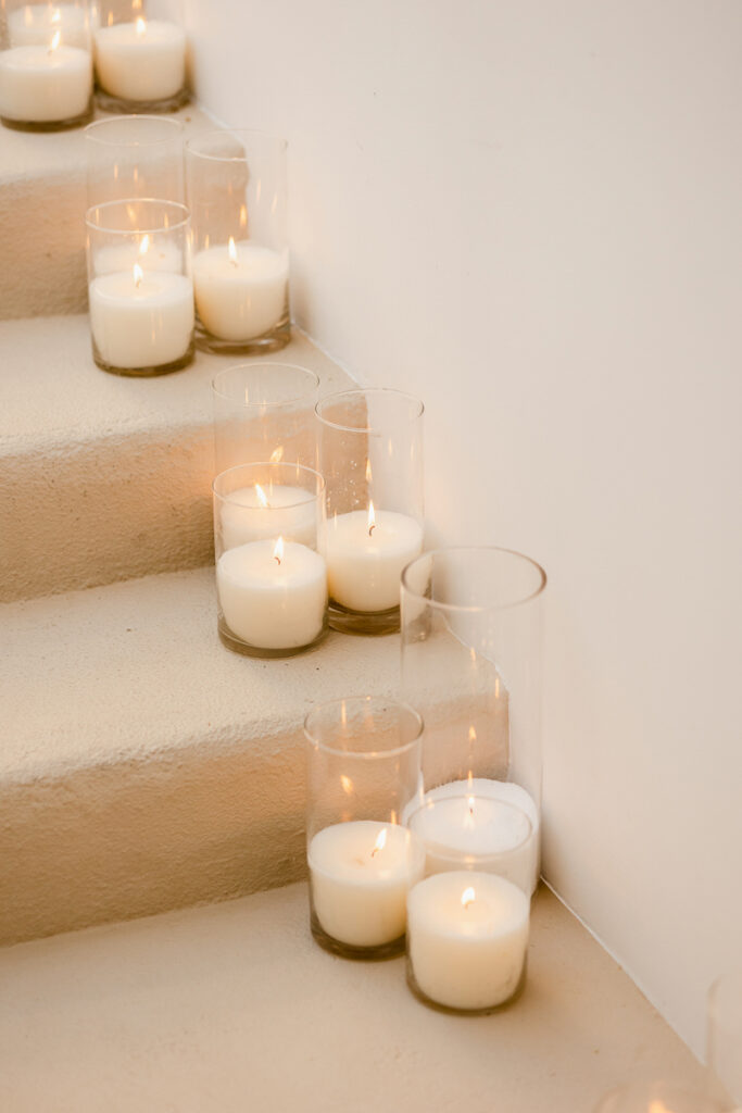 White candles in clear glass holders burning on carpeted stairs beside a white wall