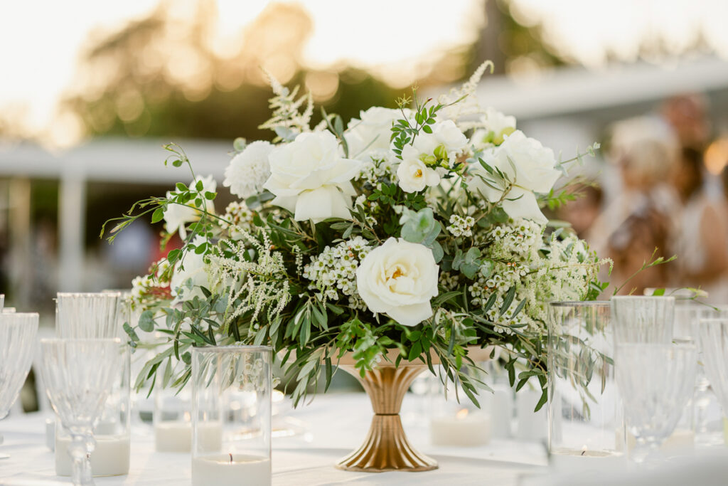 White rose and greenery floral centerpiece in a gold vase with glassware and candles on a table.
