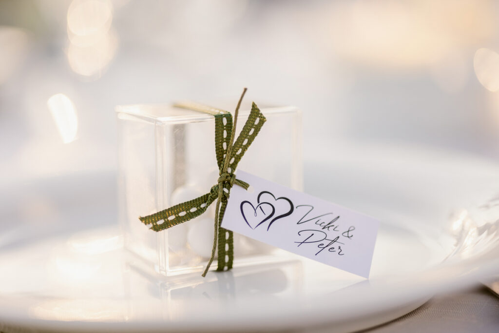Transparent gift box with olive green ribbon and tag with two hearts and names on a white plate.