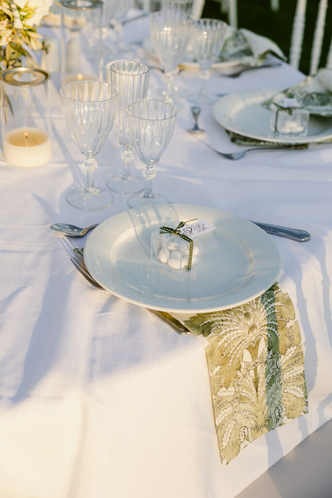 Formal table setting with white plate, clear gift box, patterned napkin, silverware, and glassware on white tablecloth.