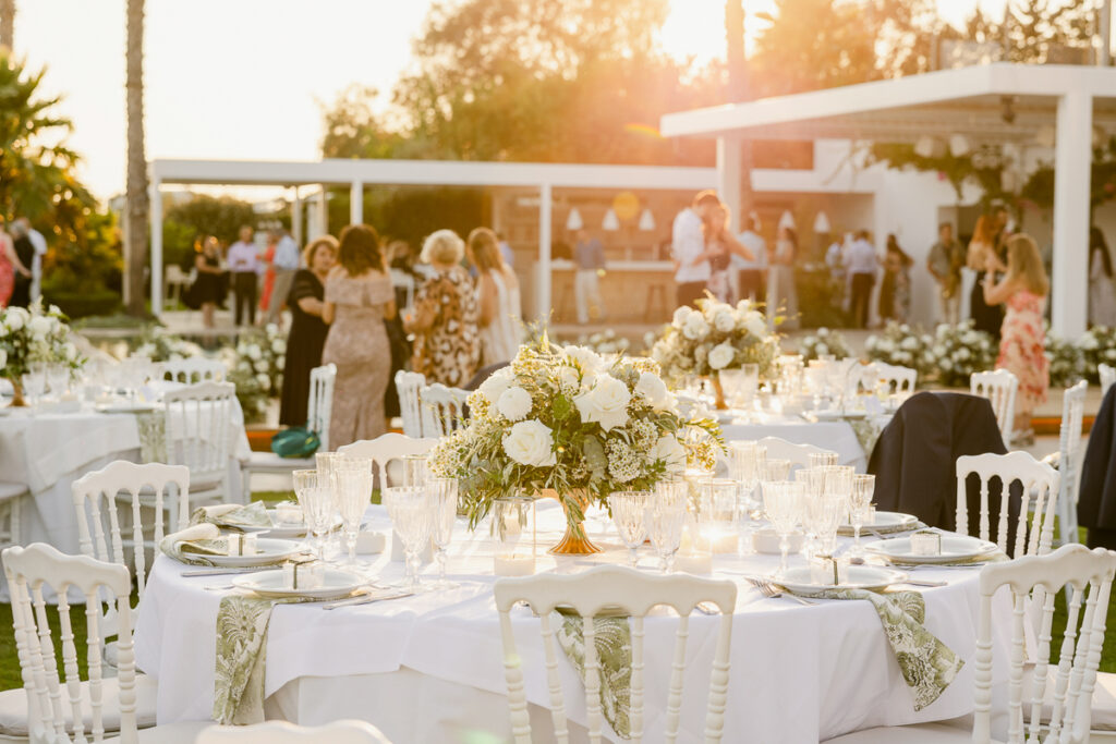 Outdoor event with white tablecloths, floral centerpieces, glassware, and guests mingling near a pavilion.