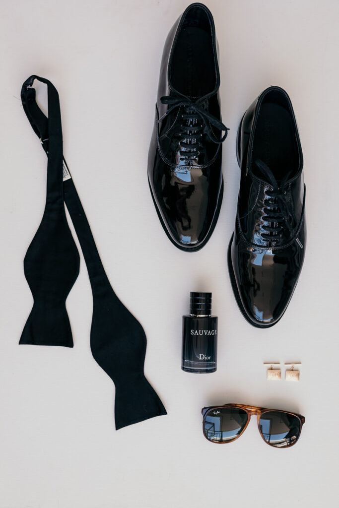 Black men's dress shoes, untied bow tie, Dior Sauvage cologne, cufflinks, and sunglasses on white surface