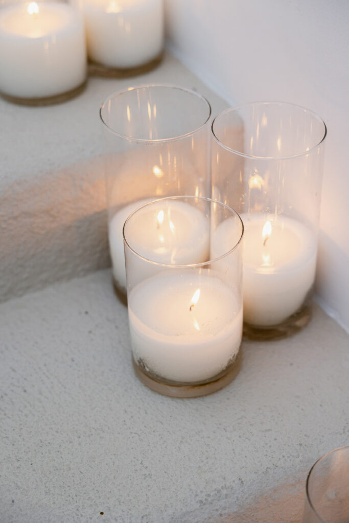 Three lit white candles in clear glass holders placed on a textured light-colored surface near a corner wall.