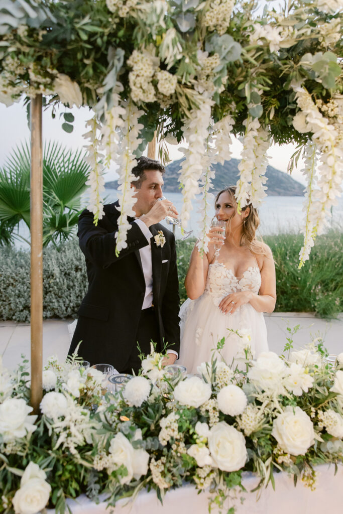 Bride and groom in wedding attire drinking at a flower-adorned outdoor table by water.