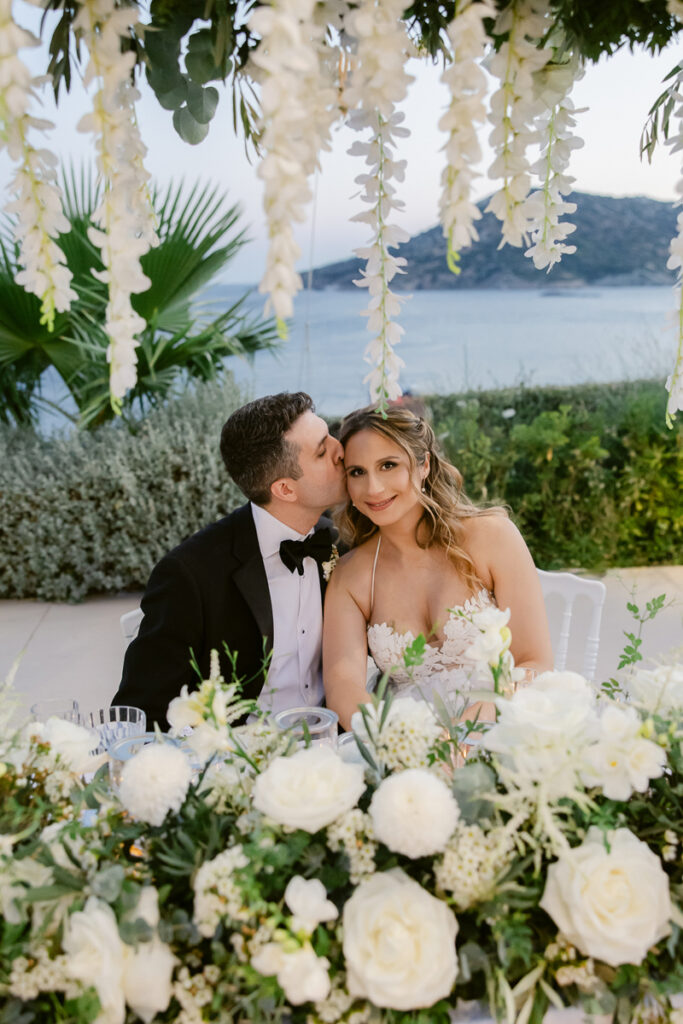 Bride and groom sit at a flower-adorned table outdoors by water with hanging white flowers above.