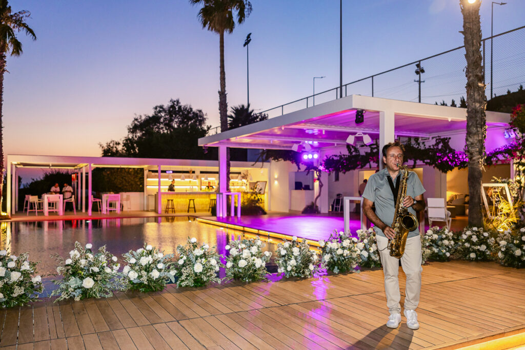 Man playing saxophone on wooden deck near pool with floral decor and illuminated bar.