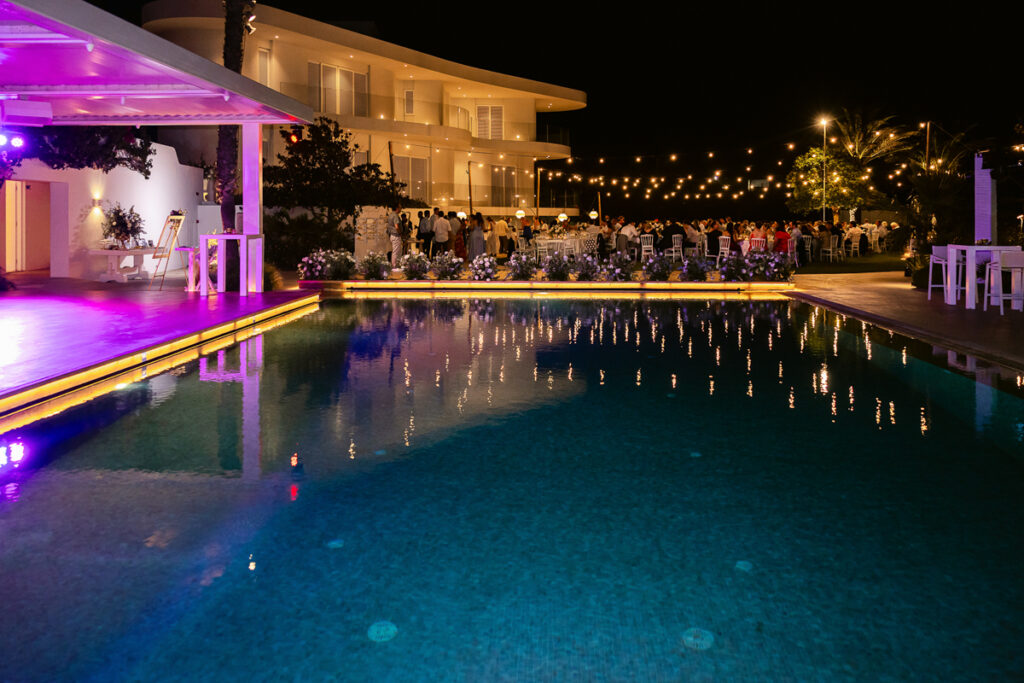 Nighttime poolside event with guests, string lights, floral decorations, and a modern building.