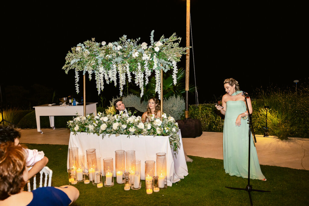 Nighttime outdoor wedding with bride and groom at floral table and woman speaking by microphone.