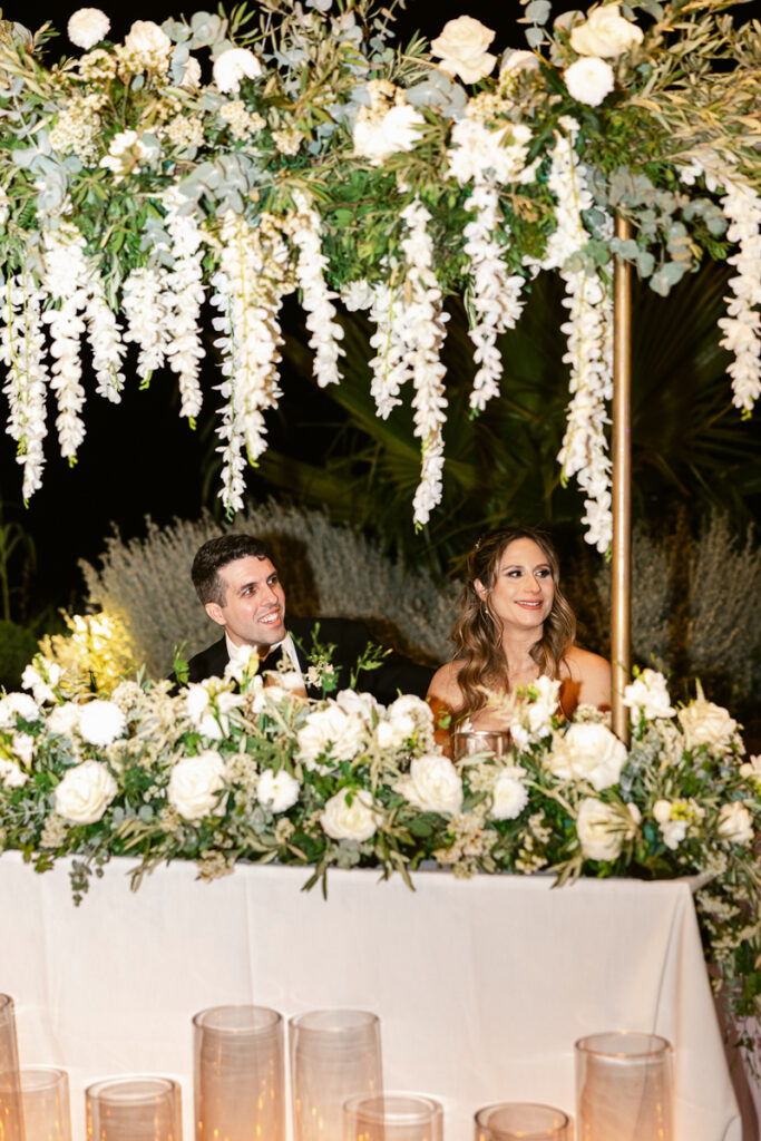 Man and woman sit behind a flower-adorned table with hanging white floral decorations at night.