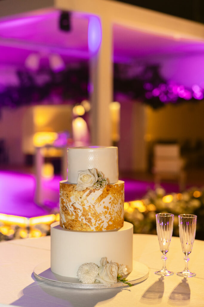 Three-tier white and gold cake with white flowers on table next to two empty champagne flutes.