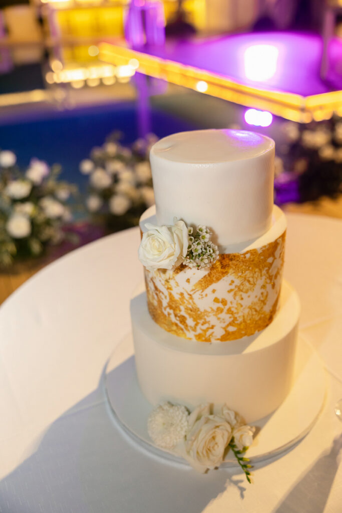 Three-tier white wedding cake with gold accents and white flowers on a round table.