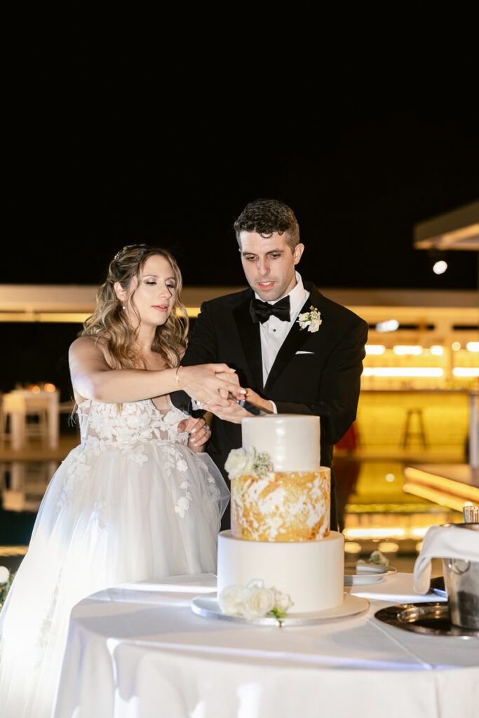 Bride and groom cutting a three-tier white and gold wedding cake at nighttime reception.