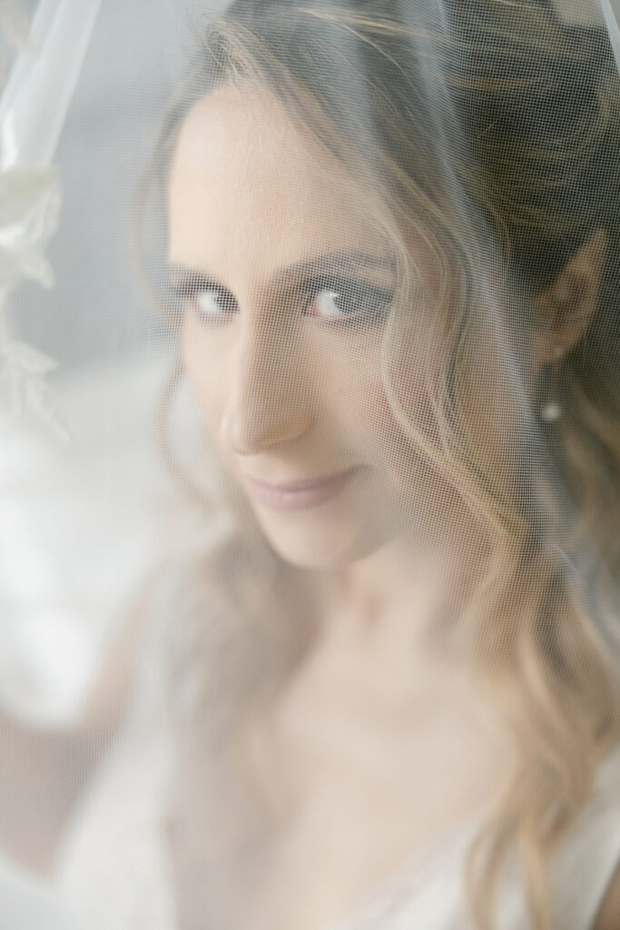 Close-up portrait of a woman with wavy hair behind a sheer bridal veil looking at the camera.