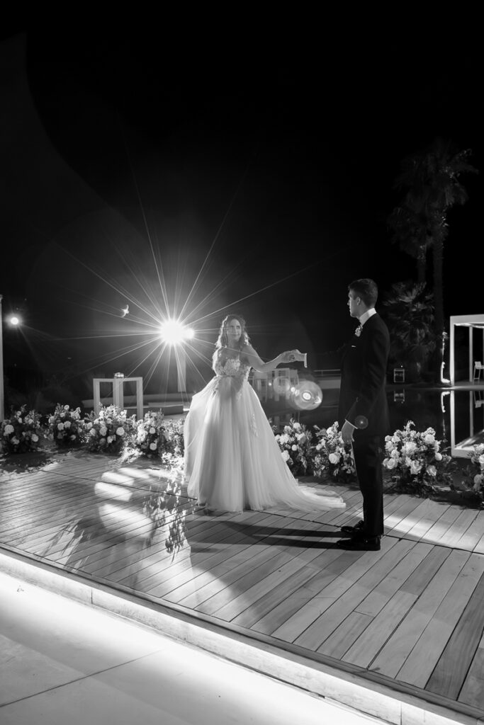 Bride in wedding gown and groom in dark suit dancing on a decorated wooden deck at night.
