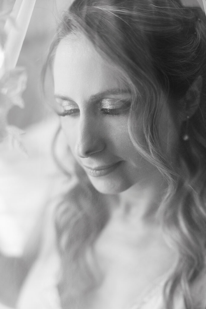 Close-up black and white photo of a woman with wavy hair behind a sheer veil with eyes closed.