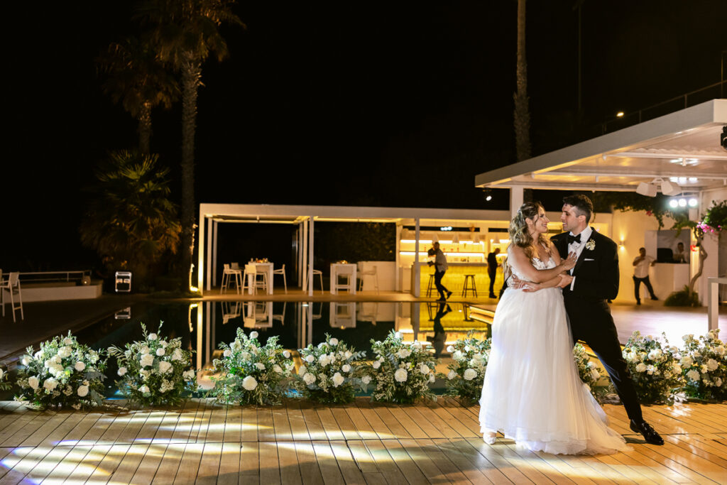 Bride and groom dancing on a wooden deck at night near floral arrangements by a pool.