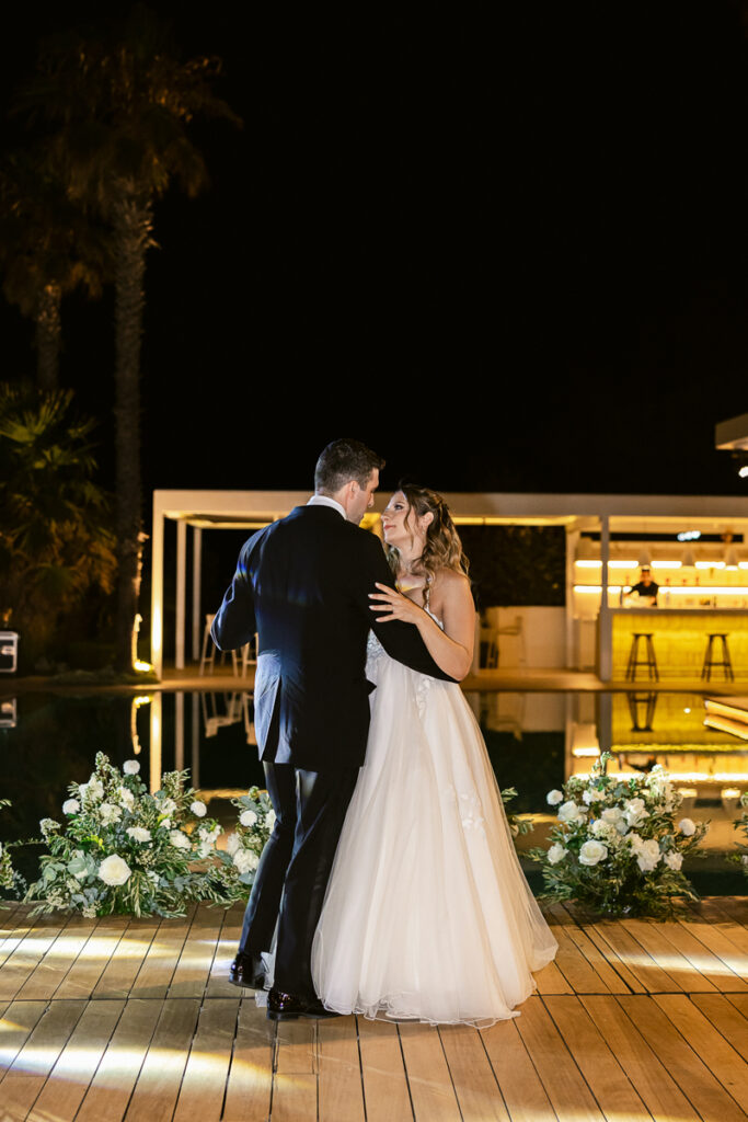 Bride and groom dancing on a wooden deck at night near a pool with floral decorations.