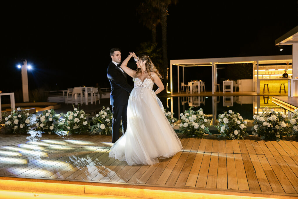 Bride and groom dancing on wooden deck by pool at night with floral arrangements and lit bar.