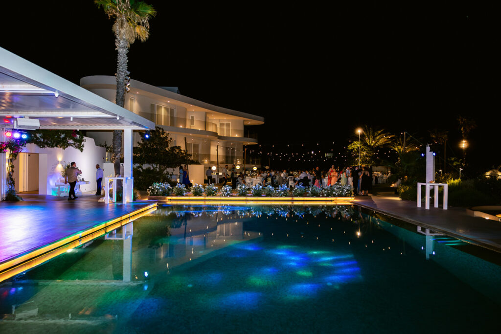 Nighttime outdoor event by illuminated pool with modern building, palm trees, and decorated seating.