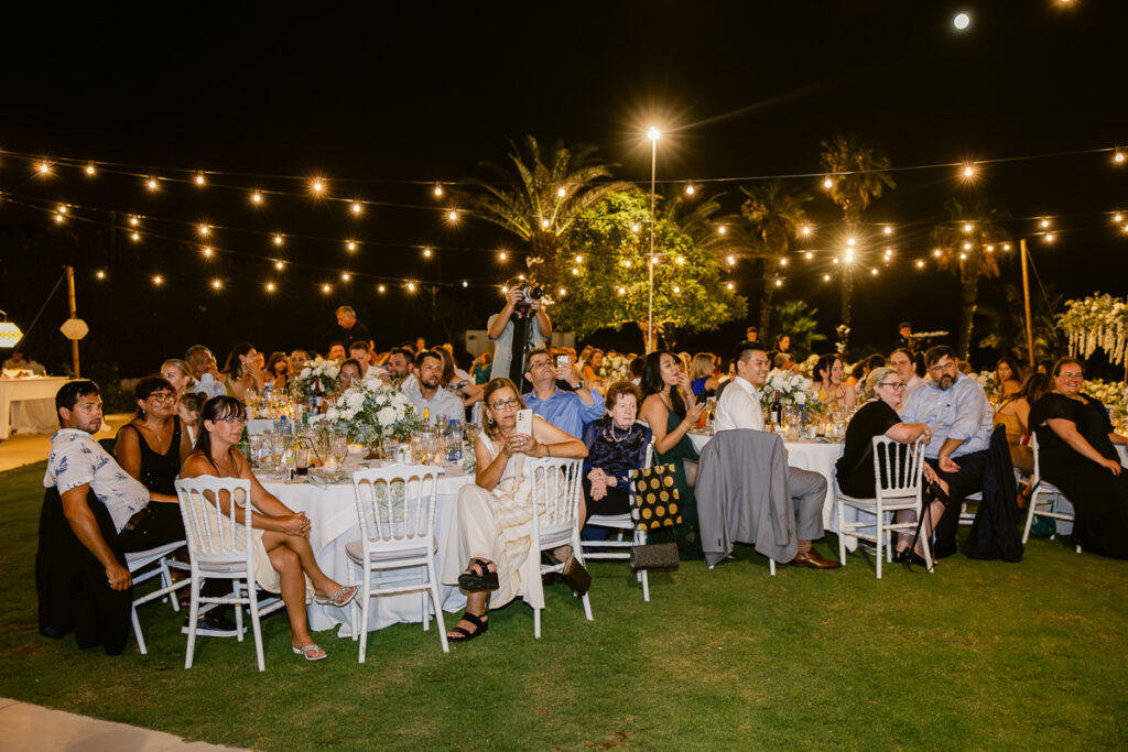 Outdoor nighttime event with guests seated at decorated round tables under string lights.