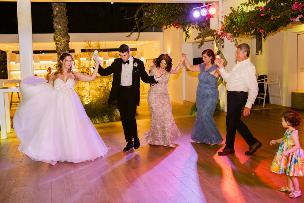 Bride, groom, and guests holding hands and dancing outdoors on wooden floor at night.
