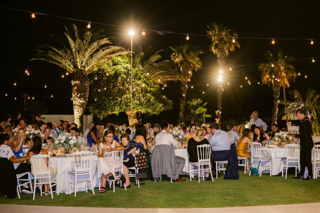 Outdoor nighttime dinner with guests seated at round tables under string lights and palm trees.