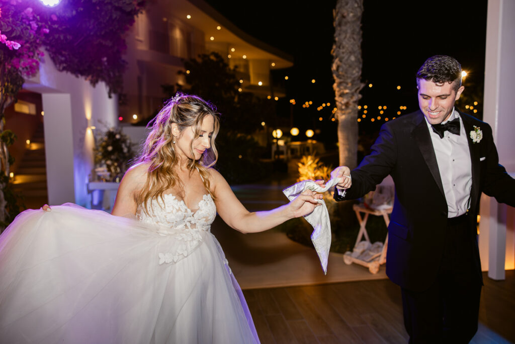 Bride in white gown and groom in tuxedo dancing outdoors at night holding a cloth.