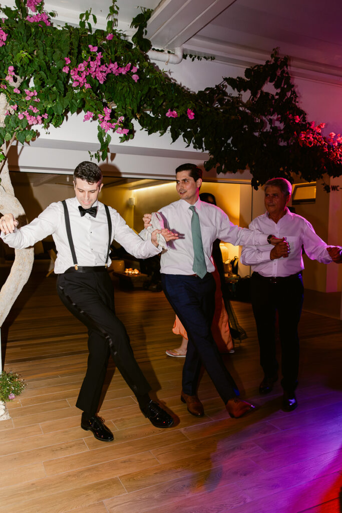 Three men in formal attire hold hands and dance indoors on a wooden floor under pink blossoms.